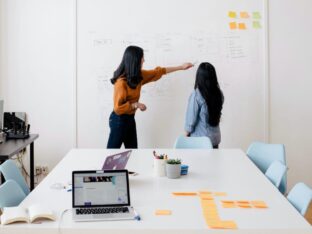 Foto de duas empreendedoras conversando em frente a um quadro branco com ideias para simbolizar o tema Plano de negócio